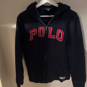 Mörkblå hoodie från Ralph Lauren - Säljer en snygg mörkblå hoodie från Ralph Lauren med stor röd POLO-text på framsidan. Den har dragkedja och en praktisk huva. Perfekt för en avslappnad stil.