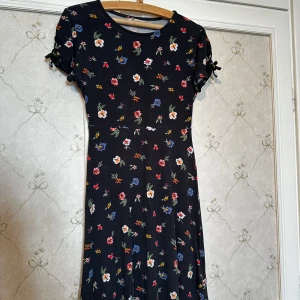 Blommig klänning i svart - Säljer en söt svart klänning med blommigt mönster i olika färger. Klänningen har korta ärmar med knytning och en rund halsringning. Perfekt för en färgglad och lekfull stil! Använd nån enstaka gång, STORLEK=146/152cl