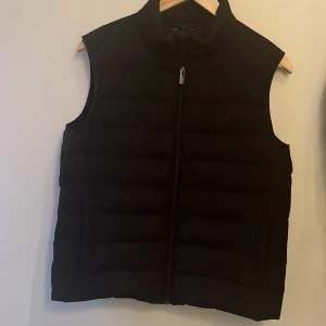 Svart dunväst från Emporio Armani - Snygg svart dunväst från Emporio Armani med dragkedja framtill. Perfekt för lager-på-lager under kyliga dagar. Klassisk design som passar till det mesta. Den har 2 st små hål en i ryggen syns på bild 5 och en på armen syns på bild 6