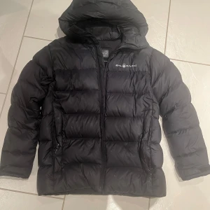 Svart dunjacka från Sail Racing - Säljer en svart dunjacka från Sail Racing. Jackan har en dragkedja framtill och en huva för extra värme. Perfekt för lite kyliga dagar. Hör av dig för minsta lilla!