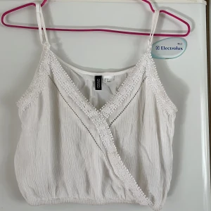 Vit omlottopp från H&M - Säljer en söt vit omlottopp från H&M med spetsdetaljer och smala axelband. Toppen har en luftig passform och är perfekt för varma dagar. Den har en fin omlottdesign framtill som ger en extra touch. Perfekt för sommaren! 🌞