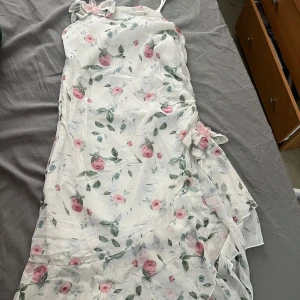 Vit blommig sommar långklänning  - Charmig vit långklänning med ett romantiskt blommönster i rosa och grönt. Klänningen har en elegant volangdetalj och en vacker blomma vid midjan. Perfekt för somriga tillfällen.                                               