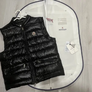 Moncler GUI - Säljer en snygg svart dunväst från Moncler med dragkedja och två fickor framtill. Västen har Monclers logotyp på bröstet och kommer med originalpåse och etikett. Perfekt för kyliga dagar! (Kan oxå bytas ut mot en colmar väst