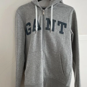 Grå hoodie från GANT - Säljer en stilren grå hoodie från GANT med dragkedja och justerbar huva. Tröjan har ett stort GANT-tryck i marinblått på framsidan. Perfekt för en avslappnad look.