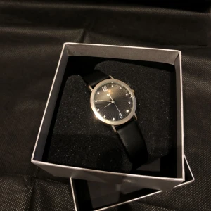 Elegant klocka med svart läderarmband - Stilren klocka med svart urtavla och silverdetaljer. Den har ett svart läderarmband och levereras i en elegant förpackning. Perfekt för en tidlös look. Nytt batteri behövs!