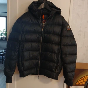 Svart Pharell Parajumpers jacka stl xxl - Säljer en svart dunjacka Pharell Parajumpers med huva och dragkedja. Jackan har ett märke på ärmen med texten 'That Others May Live'. Perfekt för kalla dagar med sin pufferdesign och långa ärmar.