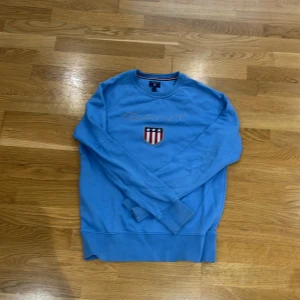 Blå sweatshirt från GANT - Säljer en snygg blå sweatshirt från GANT med broderad logga och ett emblem på bröstet. Tröjan har långa ärmar och rund halsringning. Perfekt för en avslappnad stil.