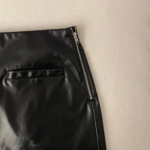 Svart skinnkjol med dragkedja - Snygg svart skinnkjol med dragkedja på sidan. Kjolen har en kort längd och stilrena fickdetaljer. Bra skick med undantag för en liten repa men påverkar inte helhetsintrycket