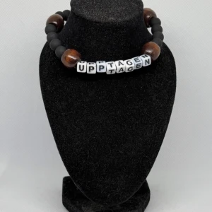 Armband med textpärlor upptagen  - Unikt armband med svarta matta pärlor och stora bruna träpärlor. Mitt på armbandet finns vita bokstavspärlor som bildar ordet 'UPPTAGEN'. Perfekt för att uttrycka din personlighet.