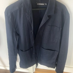 Mörkblå jacka från J.Lindeberg - Snygg mörkblå jacka från J.Lindeberg med klassisk krage och tre praktiska fickor framtill. Perfekt för en stilren look. Passar bra till både vardag och mer uppklädda tillfällen. Endast använd vid ett tillfälle alltså som ny!
