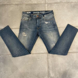 Blå jeans från Jack & Jones - Snygga blå jeans från Jack & Jones med slitna detaljer och en klassisk femficksdesign. De har en knappgylf och är perfekta för en avslappnad stil. Märkesloggan syns tydligt på insidan av midjan.