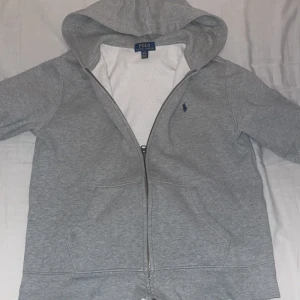 Grå hoodie från Polo Ralph Lauren - Säljer en stilren grå hoodie från Polo Ralph Lauren med dragkedja och klassisk logga på bröstet. Perfekt för en avslappnad look. Den har en mjuk insida och en praktisk huva. Storleken är 14-16 