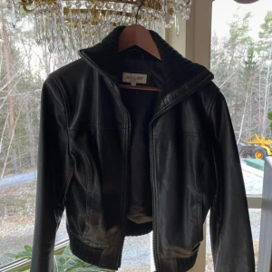 Svart skinnjacka från Hollies - Snygg svart skinnjacka från Hollies med dragkedja och ribbade detaljer. Jackan har en klassisk design med långärmade ärmar och en bekväm passform. Perfekt för att ge din outfit en cool touch. Lite nopprig i muddar/krage men går lätt att ta bort med noppmaskin! 