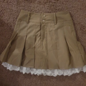 Beige kortkjol med spetsdetalj - Snygg beige kortkjol med veckad design och vit spetskant som sticker fram nedtill. Kjolen har knappar framtill och en stilren look som passar perfekt för olika tillfällen.