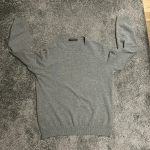 Grå ulltröja från Oscar Jacobson - Säljer en stilren grå tröja från Oscar Jacobson, tillverkad i en mjuk ullblandning. Tröjan har en klassisk design med ribbade muddar vid ärmslut och nederkant. Perfekt för kyligare dagar
