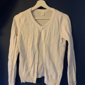 Vit kofta från H&M - Säljer en klassisk vit kofta från H&M:s Basic-kollektion. Koftan har en v-ringad design med knappar framtill och långa ärmar. Perfekt för lager-på-lager-stil eller som ett lättare ytterplagg.