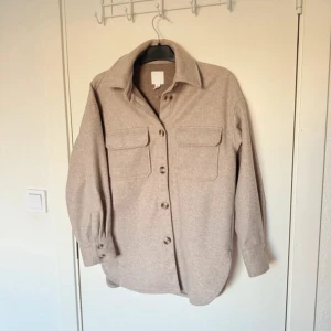Beige overshirt från H&M - Ej använd. Passar M storlek mer. Säljer en stilren beige overshirt från H&M. Jackan har en klassisk krage och knäppning framtill med stora knappar. Två praktiska bröstfickor med lock ger en cool look. Perfekt för lager-på-lager-stil under höst och vår.