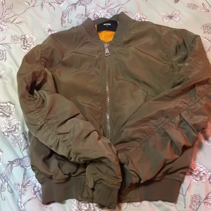 Olivgrön bomberjacka från BikBok - Snygg olivgrön bomberjacka från BikBok med dragkedja framtill och ribbade muddar. Jackan har en klassisk bomberstil med en orange insida som ger en cool kontrast. Perfekt för en avslappnad look.