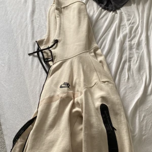 Beige hoodie från Nike - Säljer en beige hoodie från Nike med dragkedja och huva. Jackan har en svart dragkedja framtill och en extra ficka med dragkedja på ärmen. Perfekt för en sportig look.