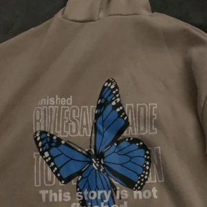 Beige hoodie med fjärilstryck - Säljer en beige hoodie med en stor blå fjäril och text på ryggen. Hoodien har en justerbar dragsko i huvan och är perfekt för en avslappnad stil.