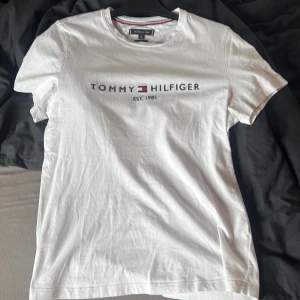 Vit t-shirt från Tommy Hilfiger - Säljer en klassisk vit t-shirt från Tommy Hilfiger med deras ikoniska logga tryckt på framsidan. T-shirten är i slim fit och har korta ärmar. Perfekt för en stilren och avslappnad look. Stl L, men passar även jättebra till dig som har M.