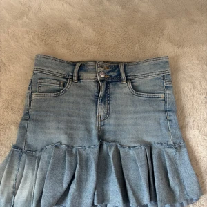 Jeanskjol med volang - Trendig jeanskjol i ljusblå denim med volangdetalj nedtill. Kjolen har klassiska femficksdetaljer och stängs med knapp och dragkedja framtill. 