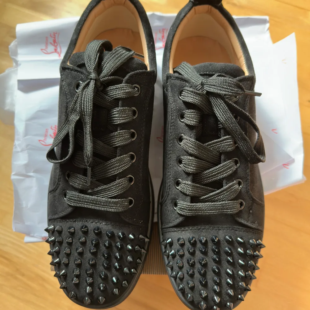 Snygga svarta sneakers från Christian Louboutin med nitar på tån och klassisk röd sula. Skorna har snörning och är tillverkade i mocka. Perfekta för att ge din outfit en edgy touch.. Kengät.