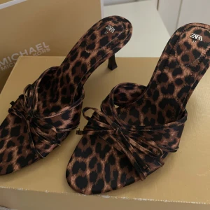 Leopardmönstrade klackskor från Zara - Snygga leopardmönstrade klackskor från Zara med öppen tå och elegant rosettdetalj. Perfekta för att ge en djärv touch till din outfit. Klackarna är smala och ger en stilren look.