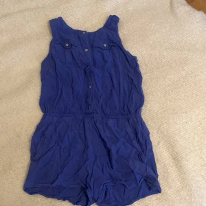 Blå ärmlös klänning med shorts  - Säljer en snygg blå ärmlös klänning med shorts  med knappar framtill. Perfekt för varma dagar och enkel att styla med accessoarer. Den har en elastisk midja för en bekväm passform. Den är i bra skick. Inga hål. Inga fläckar 