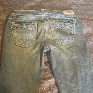 Blå jeans från True Religion - Säljer ett par klassiska blå jeans från True Religion med snygga kontrastsömmar och ikoniska bakfickor. Jeansen har en låg midja och en bootcut-stil som ger en avslappnad look. Perfekta för dig som vill ha en tidlös stil med en touch av edge.