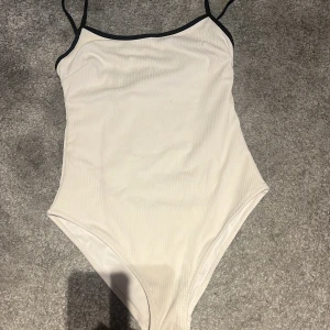 Vit ribbad baddräkt från H&M - Säljer en stilren vit baddräkt från H&M med svarta tunna axelband och svarta kanter. Baddräkten är ribbad och har en klassisk, enkel design som passar perfekt till stranden eller poolen.