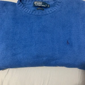 Blå stickad tröja från Polo Ralph Lauren - Säljer en klassisk blå stickad tröja från Polo Ralph Lauren i 100% bomull. Tröjan har rund halsringning, långa ärmar och den ikoniska broderade loggan på bröstet. Perfekt för en stilren och avslappnad look.