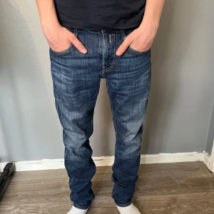 Blå Replay jeansbyxor - Säljer ett par klassiska blå jeans från Replay med snygga slitningar och kontrastsömmar. Byxorna har fem fickor, dragkedja och knapp framtill samt Replay-logga på bakfickan och läderpatch i midjan.
