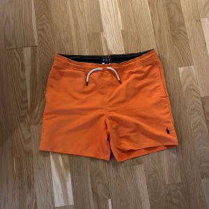 Orange badshorts från Polo Ralph Lauren - Snygga orange badshorts från Polo Ralph Lauren med vit snörning i midjan och klassisk broderad logga på benet. Shortsen har en bakficka med knapp och Polo Ralph Lauren Swimwear-märke nypris är 1000kr. Pris kan diskuteras 