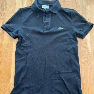Pikétröjor från Lacoste stl S - Klassisk svart och ljusgrön pikétröja från Lacoste med krage och knappar framtill. Tröjan har korta ärmar och den ikoniska gröna krokodilloggan på bröstet. Slim fit-modell som passar perfekt till många tillfällen.