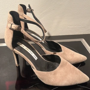 Beige mocka pumps med ankelrem från & Other Stories - Säljer ett par snygga beige pumps från & Other Stories i mocka med spetsig tå och smal klack. Skorna har en elegant ankelrem med spänne som ger extra stöd och stil. Perfekta för dig som vill ha en klassisk men trendig look.