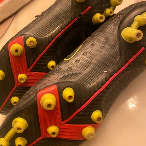 Nike Mercurial fotbollsskor med dobbar - Säljer ett par svarta Nike Mercurial fotbollsskor med gula och röda detaljer. Skorna har snörning, hög krage och gula dobbar för bra grepp på planen. Perfekta för dig som vill ha lätta och snabba skor till matchen.