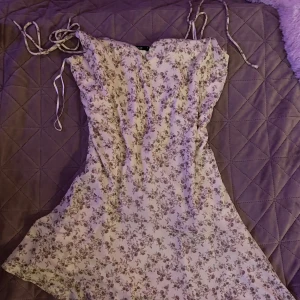 Blommig sommarklänning med smala axelband - Söt ljus klänning med små blommor i lila och grönt. Klänningen har smala knytbara axelband och en lätt, luftig passform. Perfekt för varma dagar och har en feminin, romantisk stil. De speciella med klänningen är att den åker lite snett vid benet🩵