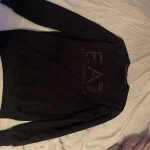 Svart sweatshirt från Emporio Armani EA7 - Säljer en svart sweatshirt från Emporio Armani EA7 med diskret logga på bröstet. Tröjan har rund halsringning och långa ärmar. Perfekt för en stilren och enkel look. Passar utmärkt till Storlek M