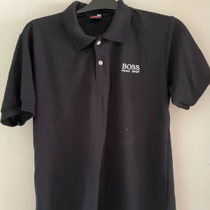 Svart pikétröja från Boss - Säljer en klassisk svart pikétröja från Boss med vit broderad logga på bröstet och två knappar vid kragen. Perfekt för en stilren och sportig look.