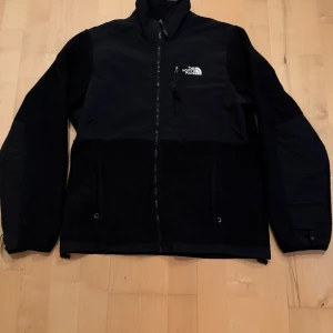 Svart fleecejacka från The North Face - Säljer en svart fleecejacka från The North Face med dragkedja framtill och logga på bröstet och ryggen. Jackan har hög krage och en bröstficka med dragkedja. Perfekt för kyliga dagar och enkel att matcha. Finns liten skavank på baksidan. Står dam L men passar också men S.