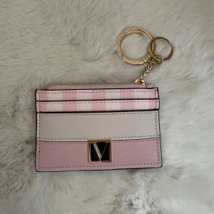 Plånbok från Victoria Secret - Supersöt plånbok från Victoria Secret i rosa och vitt med rutigt mönster upptill. Har guldfärgad nyckelring och ett V-emblem framtill. Perfekt för kort och mynt, smidig att ha med sig överallt. Jag köpte fel design, säljer denna för att köpa en ny. 