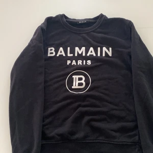 Svart sweatshirt från Balmain Paris - Säljer en svart sweatshirt från Balmain Paris med vit logotyptryck framtill. Tröjan har rund halsringning och långa ärmar. Perfekt för en stilren och trendig look.