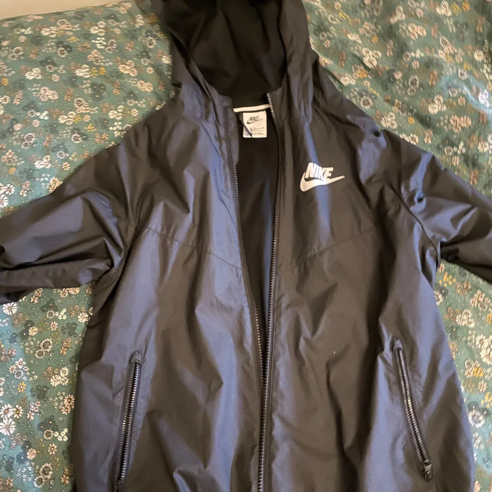 Svart vindjacka från Nike med huva och dragkedja framtill. Jackan har två sidofickor med dragkedja och en vit Nike-logga på bröstet. Perfekt för blåsiga dagar och enkel att matcha med det mesta. Ögglan för att hänga upp jackan har gått sönder som man ser på bild 3. Storlek 158-170cm, XL i barn, ungefär som xs i vuxen storlek. . Takit.