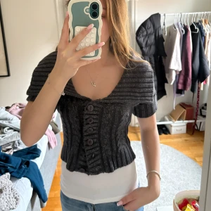 Mörkgrå stickad kofta med knappar - Säljer en mörkgrå stickad kofta med kort passform och stora dekorativa knappar framtill. Koftan har bred krage och är kortärmad, perfekt att bära över ett linne eller t-shirt. Materialet är en blandning av akryl, polyester och metallisk tråd för en lätt glittrig effekt.