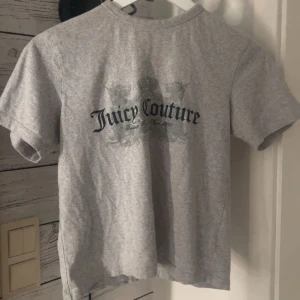 Grå t-shirt från Juicy Couture - Grå t-shirt från Juicy Couture med tryck på ryggen. Klassisk passform och rund halsringning. Perfekt för en avslappnad stil och enkel att matcha med jeans eller shorts.