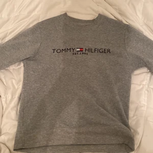 Grå sweatshirt från Tommy Hilfiger - Säljer en klassisk grå sweatshirt från Tommy Hilfiger med logga och flagga broderad framtill. Tröjan har rund halsringning och långa ärmar. Perfekt för en avslappnad och stilren look.