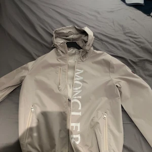 Beige vindjacka från Moncler med huva - Säljer en beige vindjacka från Moncler med huva och dragkedja. Jackan har två fickor med dragkedja framtill, stor Moncler-logga längs dragkedjan och en broderad logotyp på ärmen. Perfekt för blåsiga dagar.