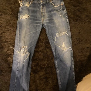 Levi's 501 slitna jeans med hål - Säljer ett par klassiska blå Levi's 501 jeans med slitningar och stora hål på benen för en riktigt cool och avslappnad look. Rak passform och normal midja. Perfekta för dig som gillar vintage och streetstyle.