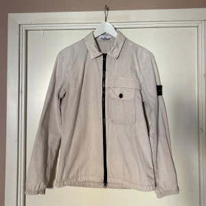 Beige jacka från Stone Island - Säljer en stilren beige jacka från Stone Island. Jackan har en dragkedja framtill och en bröstficka med knapp. Den har långa ärmar och en klassisk krage. Perfekt för en avslappnad look. 